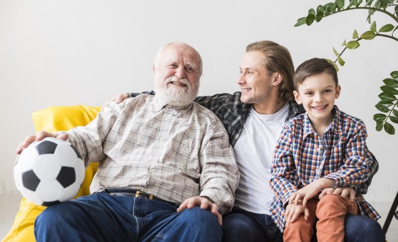 Três gerações juntas: senhor de idade, sentado ao lado de homem mais jovem, que está com uma criança em seu colo