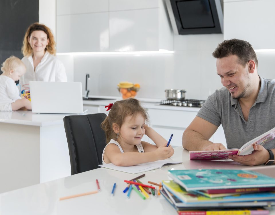 Homem lendo com filha, sentados na cozinha de casa, enquanto ao fundo está uma mulher com um bebê e um notebook em frente