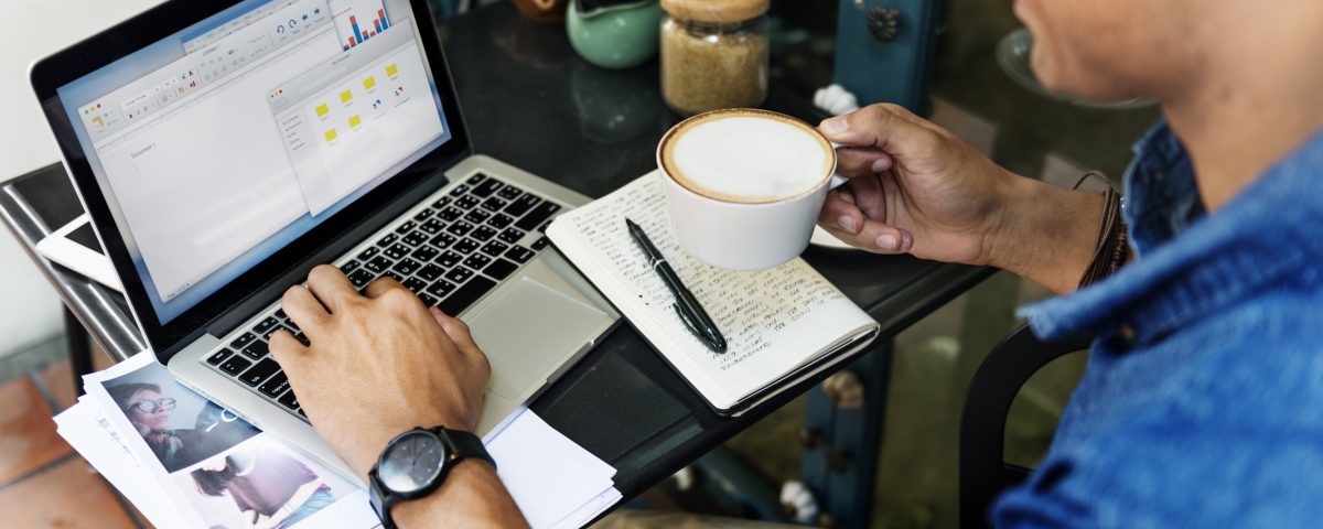 Homem sentado, trabalhando com notebook e caderno, e tomando um café