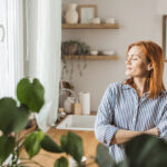Mulher, em pé, em frente de janela, com olhos fechados, pensativa e com rosto expressando calma e felicidade