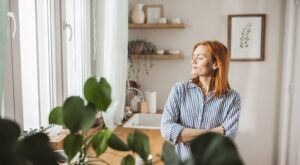 Mulher, em pé, em frente de janela, com olhos fechados, pensativa e com rosto expressando calma e felicidade